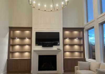 A modern living room with a beige sectional sofa, a large circular chandelier, built-in shelves with lights, a fireplace, and a wall-mounted TV, with large windows on the right.